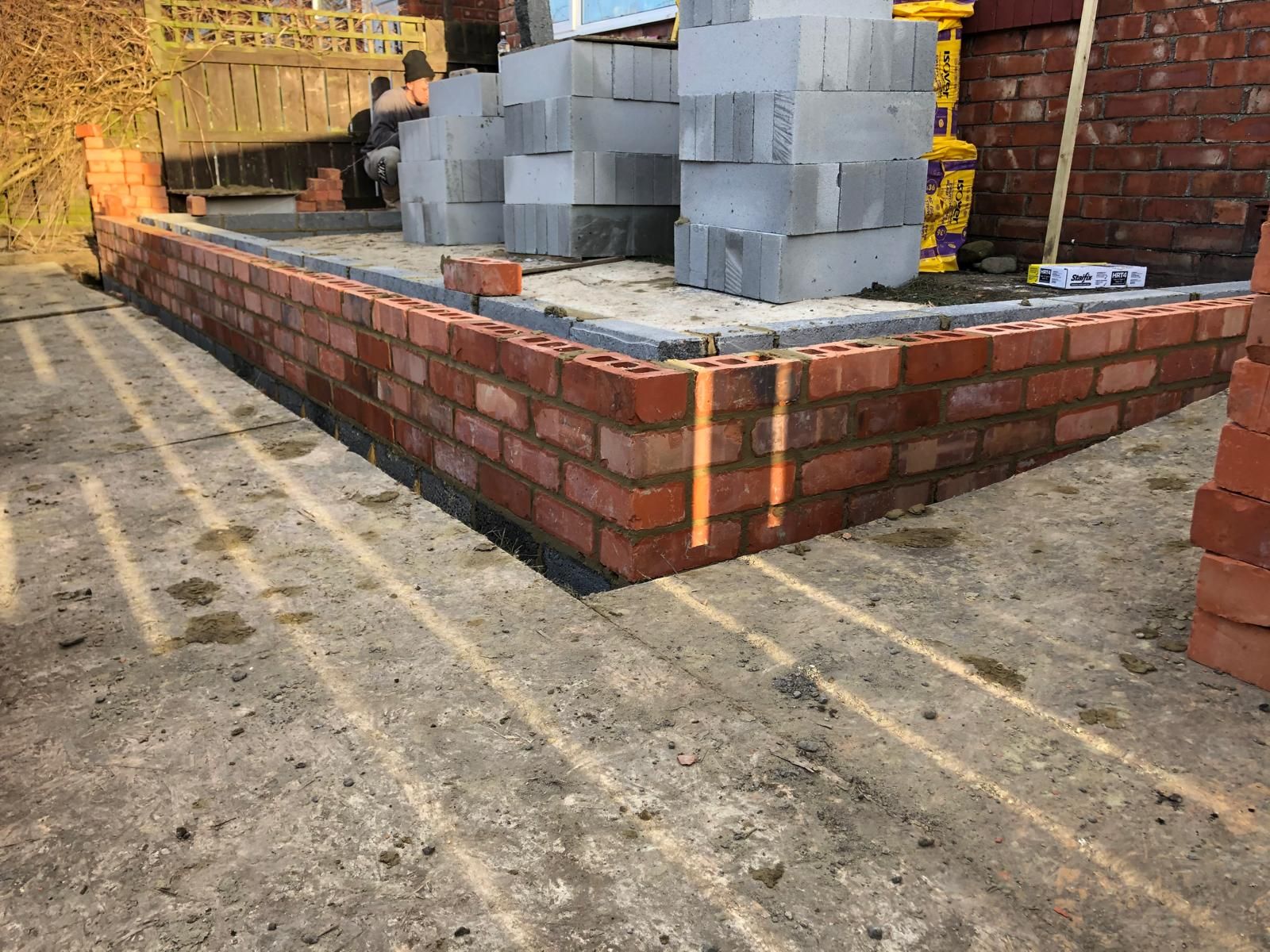 Neat new red-brick footings forming a right-angled corner on a concrete oversite with blocks and brick packs stacked behind
