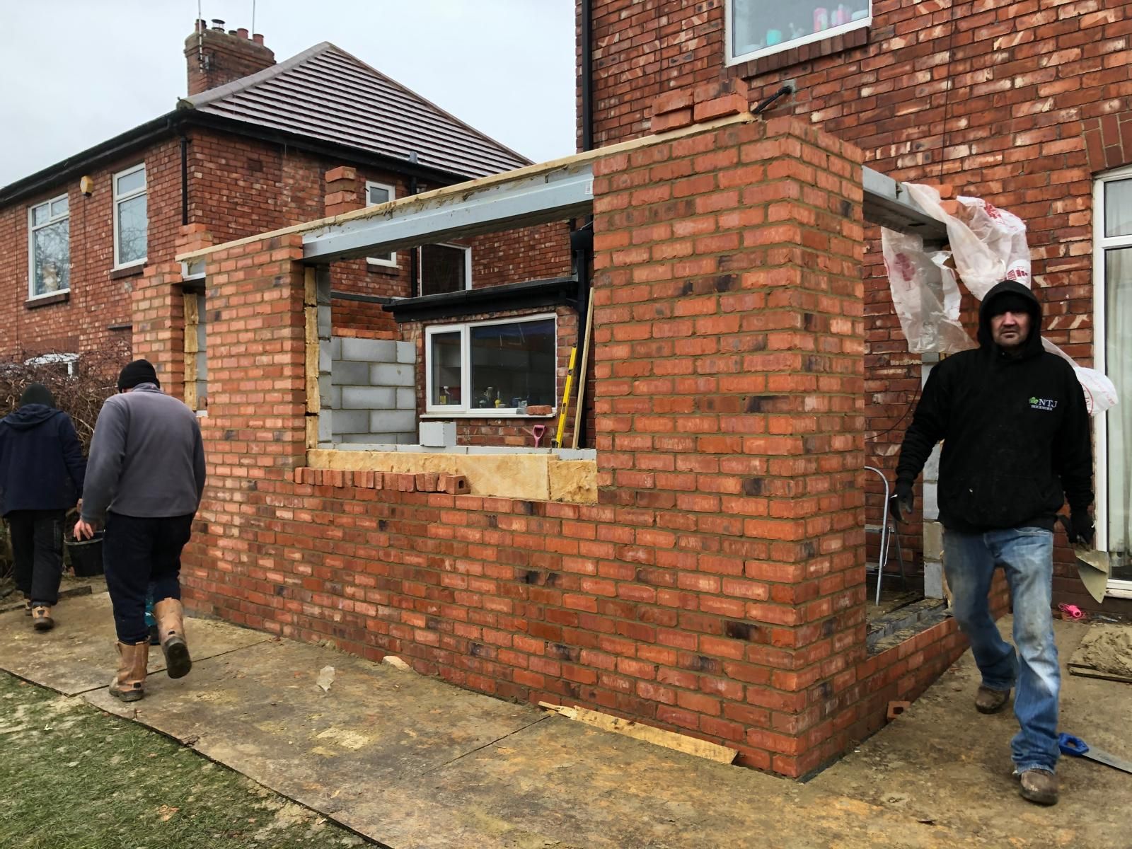 Builders forming a rear extension opening, red-brick skin built up around a structural steel beam with the original house behind