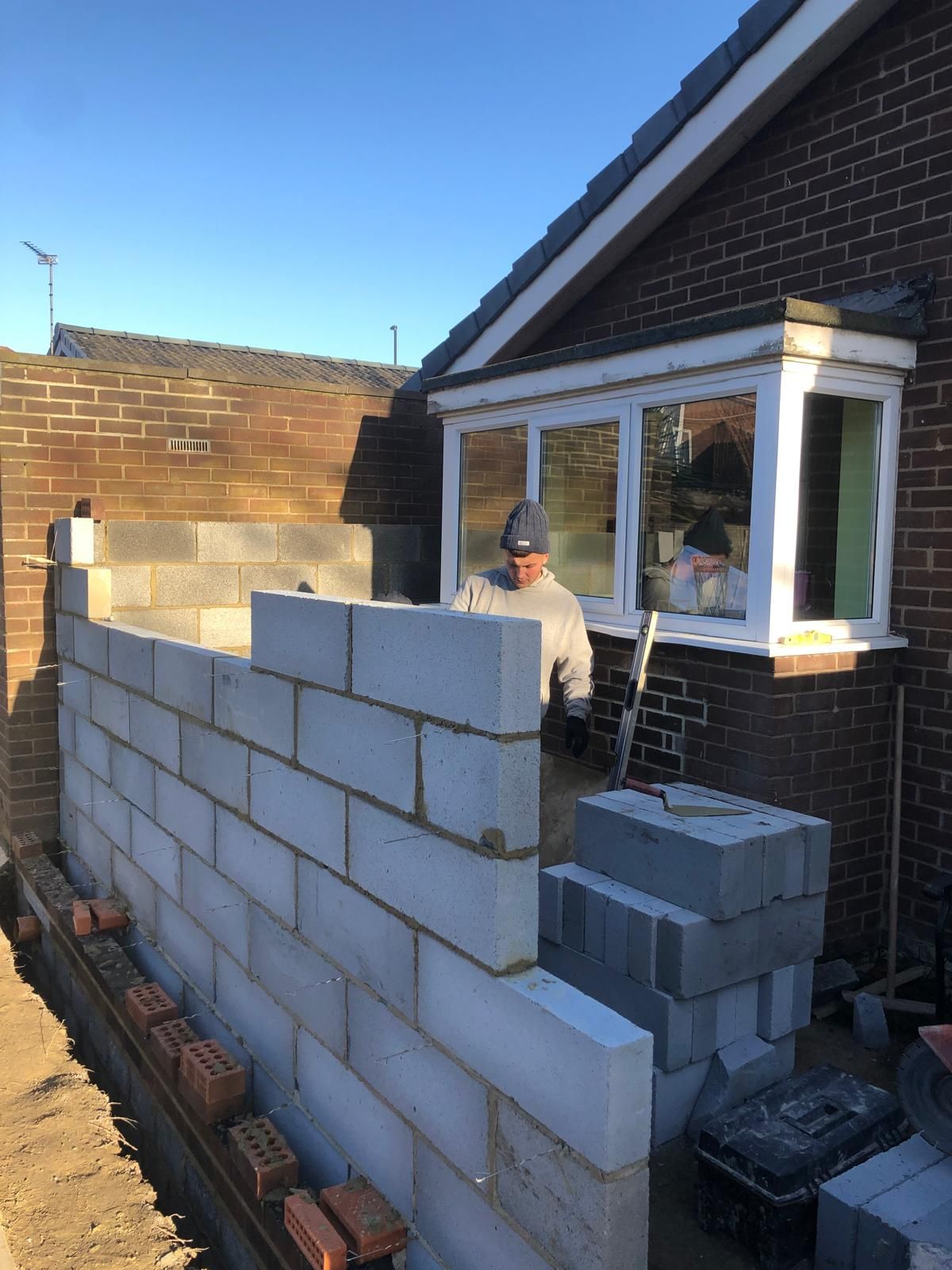 Bricklayer building a block garden wall with a red-brick pier against a bungalow under a clear blue sky