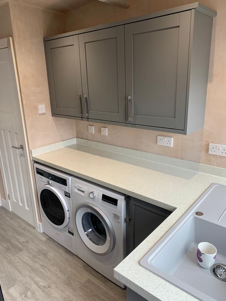 Grey shaker utility room with wall cabinets, white speckled worktop, integrated washing machine and tumble dryer and a granite-effect sink