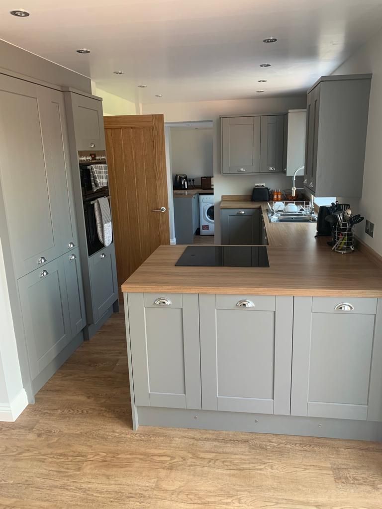 Light grey-green shaker kitchen with oak-effect worktops, cup handles, induction hob and oak internal door through to a utility area
