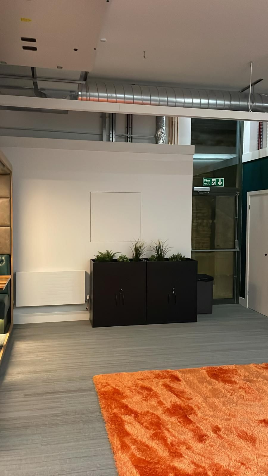 Office breakout area with black planter units, exposed silver ductwork overhead and an orange rug on grey flooring