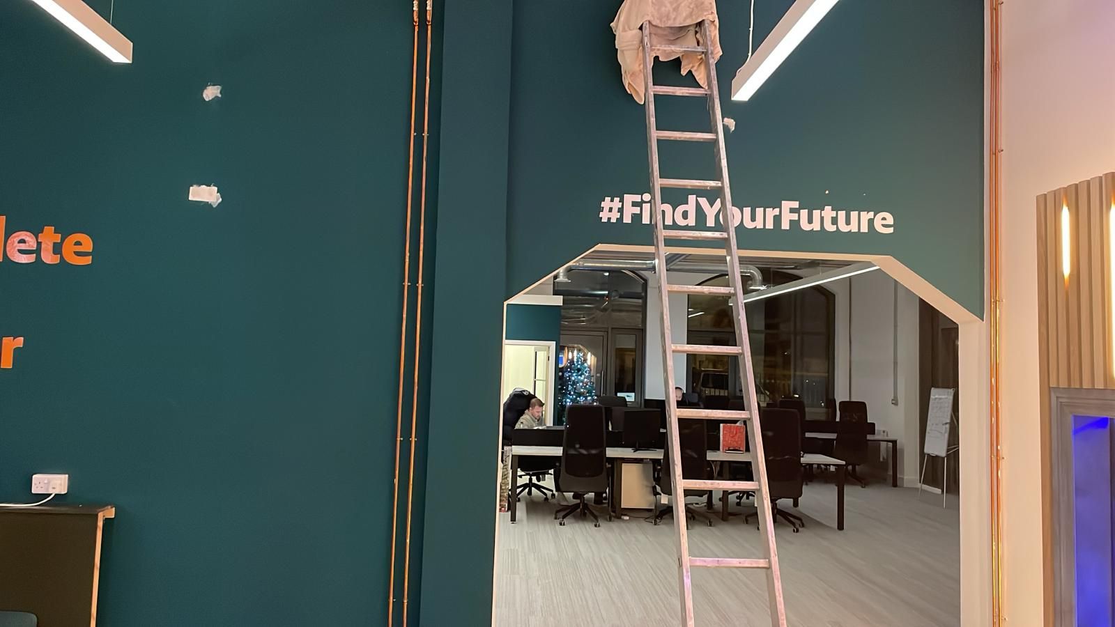 Mid-fit-out shot of the teal office with a timber ladder, exposed copper pipework and "#FindYourFuture" wall vinyl, staff working at desks behind