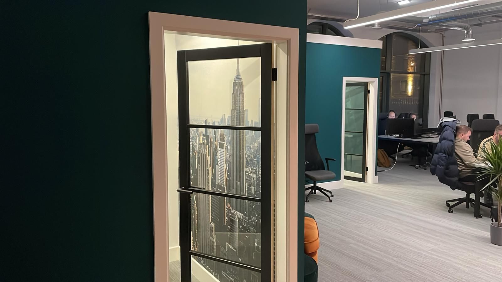 Black-framed glazed internal doors with a New York skyline graphic set into teal walls, looking through to the open-plan desks
