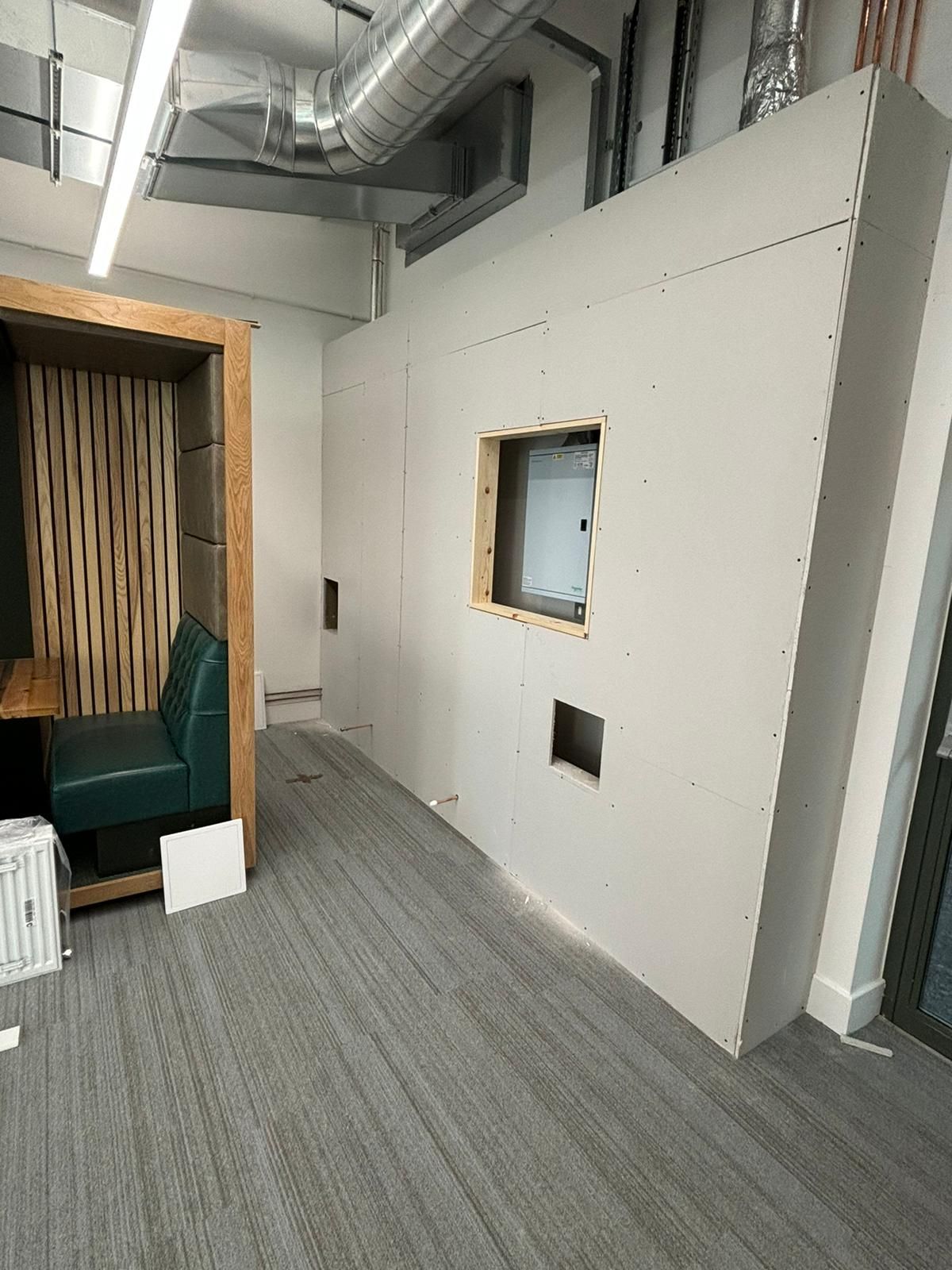 Meeting-room pod under construction with plasterboard walls, a framed serving hatch opening and an oak-clad booth seat to the side