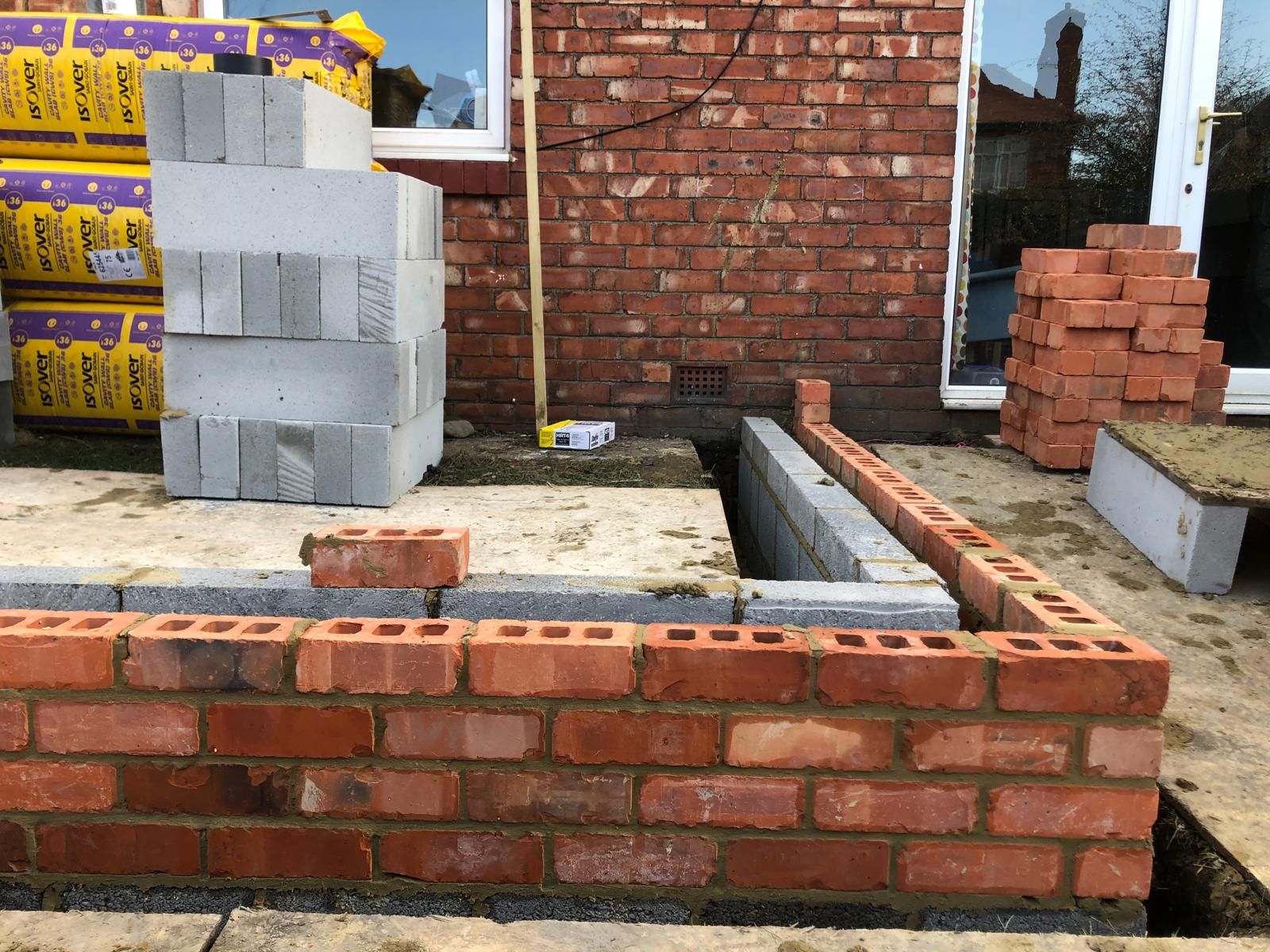 Strip foundations dug along the rear of a bungalow with concrete bases laid and a black bench against the existing brickwork